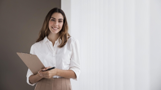 Compliance: imagem mostra uma mulher sorrindo com camisa branca e saia caramelo segurando uma prancheta e uma caneta.