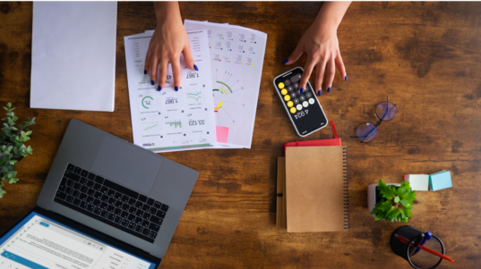 imagem mostra a mão de uma pessoa trabalhando com uma calculadora, checando números em papéis sobre a mesa e no notebook.