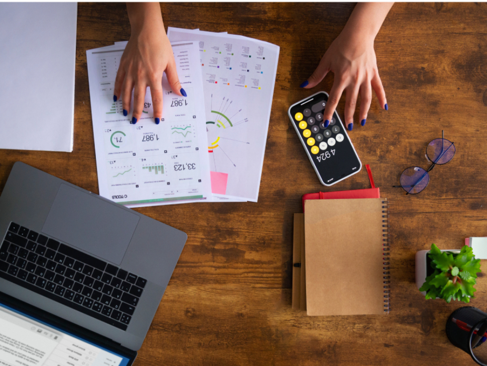 imagem mostra a mão de uma pessoa trabalhando com uma calculadora, checando números em papéis sobre a mesa e no notebook.