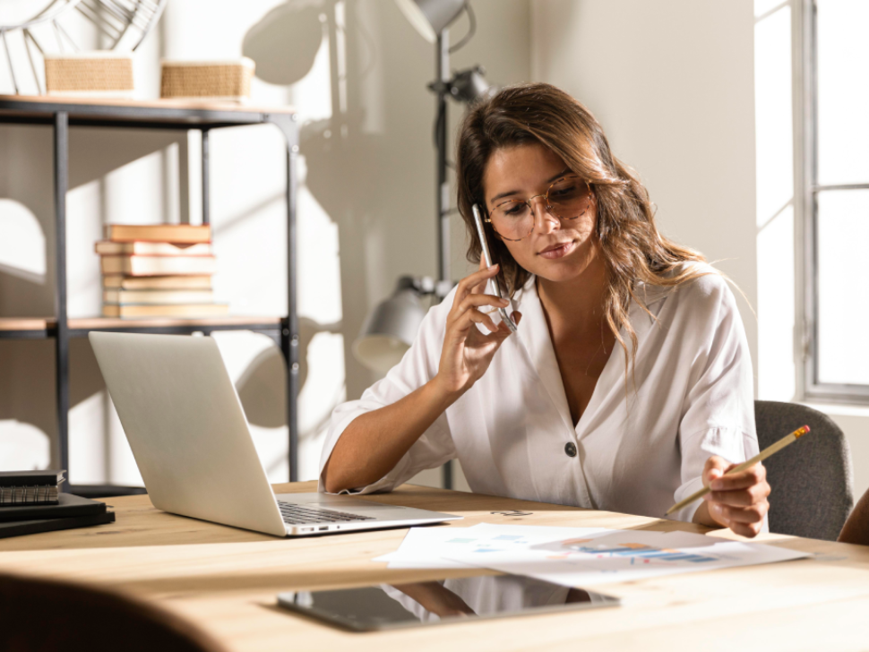 imagem mostra mulher trabalhando em sua mesa com o notebook à frente e falando ao celular.