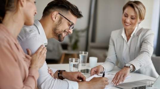 imagen muestra 3 personas sonriendo en una mesa. El hogar está firmando un papel.