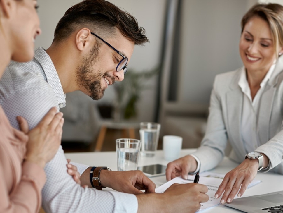 imagen muestra 3 personas sonriendo en una mesa. El hogar está firmando un papel.