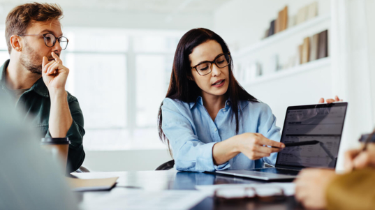 imagem mostra uma mulher apontando para a tela do notebook com relatórios financeiros e um homem analisando os dados logo atrás.