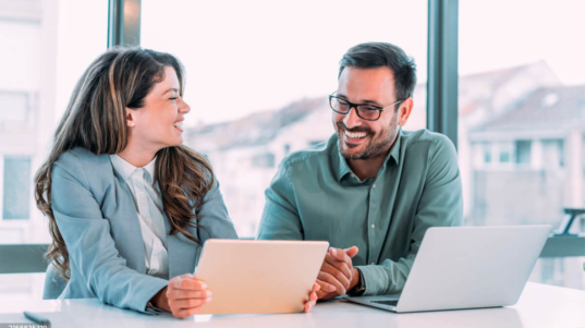 imagem mostra uma mulher e um homem sorrindo em frente a uma tela de tablet e uma tela de notebook, felizes por terem um melhor controle e otimização de custos e resultados de empresas.