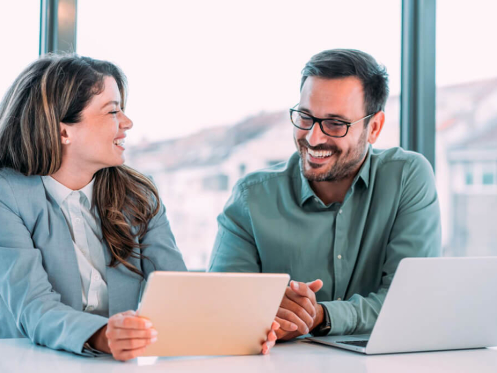 imagem mostra uma mulher e um homem sorrindo em frente a uma tela de tablet e uma tela de notebook, felizes por terem um melhor controle e otimização de custos e resultados de empresas.