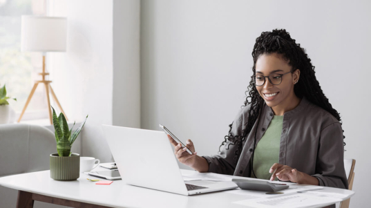 imagem mostra uma mulher sorrindo, segurando um celular e satisfeita por ter conseguido fazer um fechamento eficaz para a empresa dela. Na mesa, tem um notebook e uma calculadora.