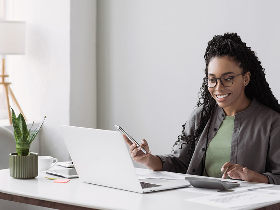 imagem mostra uma mulher sorrindo, segurando um celular e satisfeita por ter conseguido fazer um fechamento eficaz para a empresa dela. Na mesa, tem um notebook e uma calculadora.