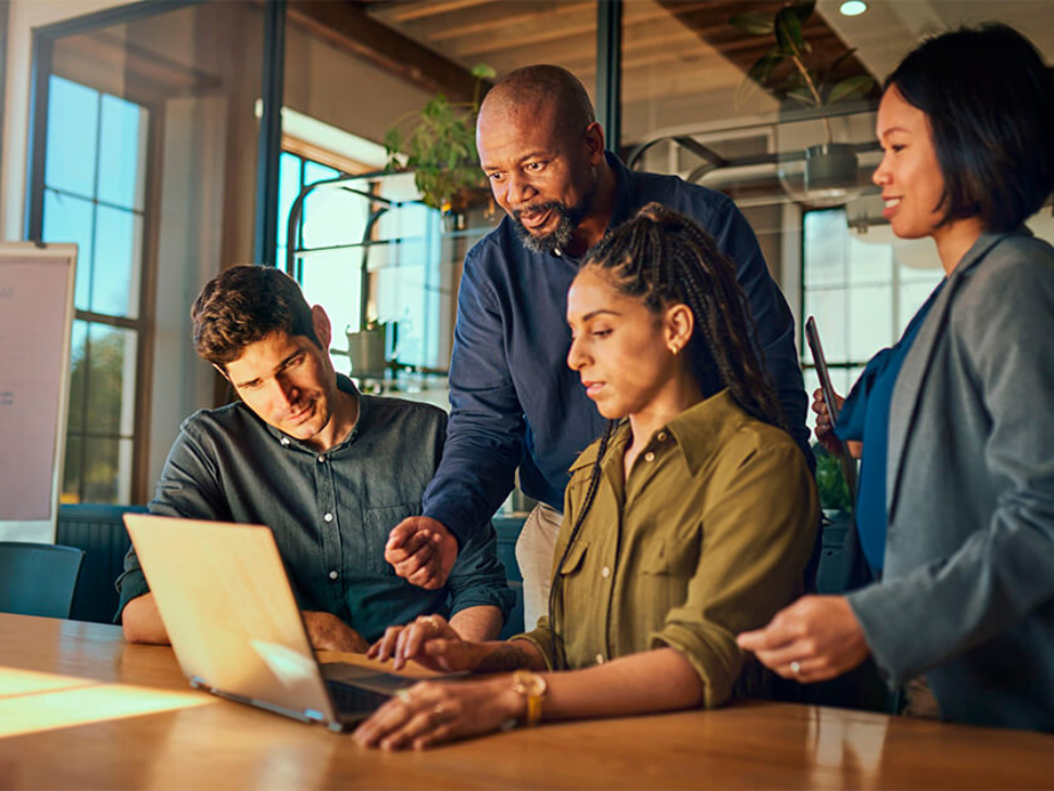 imagem mostra 4 pessoas (2 homens e 2 mulheres) analisando a tela de um laptop em um escritório.