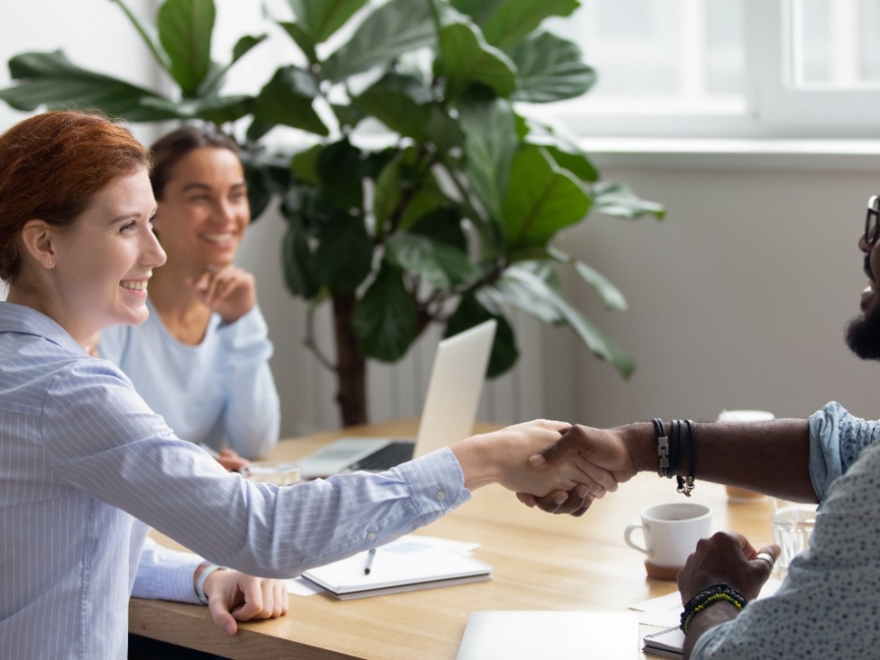 imagem mostra pessoas sentadas em uma mesa e duas pessoas apertando as mãos em sinal de acordo.