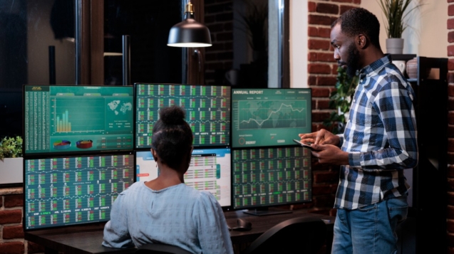 imagem mostra duas pessoas em frente a várias telas que acompanham o mercado financeiro.