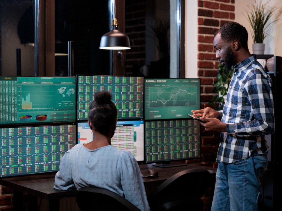 imagem mostra duas pessoas em frente a várias telas que acompanham o mercado financeiro.