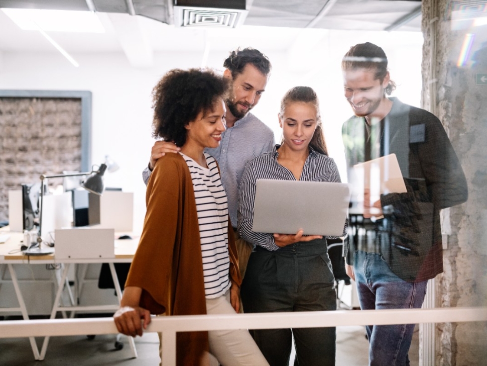 imagem mostra 4 pessoas em pé sorrindo e reunidas em volta de um notebook no escritório