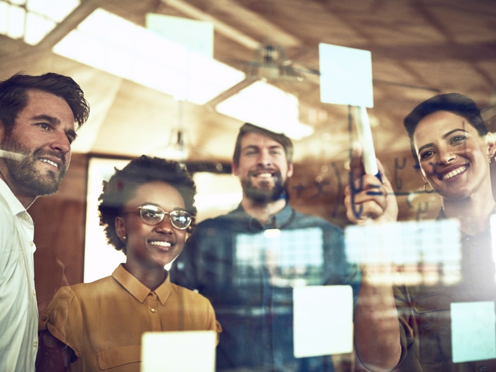 imagem mostra 4 pessoas sorrindo em um escritório colando post-its em um mural.