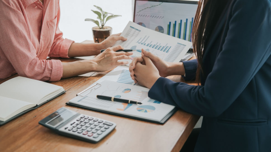 Imagem mostra duas pessoas analisando planilhas e relatórios financeiros com uma calculadora ao lado.